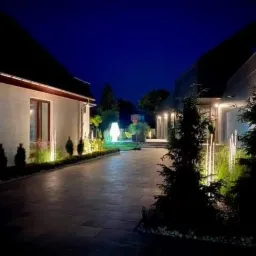 Oświetlona posesja nocą: nowoczesny dom z oświetleniem elewacyjnym i ogrodowym, widoczne lampy wzdłuż ścieżki i podświetlone krzewy.