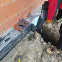 Pomiar rezystancji uziomu przy budynku z cegły, z użyciem miernika i wiertarki udarowej Bosch, widoczne narzędzia i wykop.