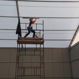 ELPA Instalacje Elektryczne - Elektryk na rusztowaniu montuje oświetlenie wewnątrz hali z metalową konstrukcją i przezroczystym dachem.