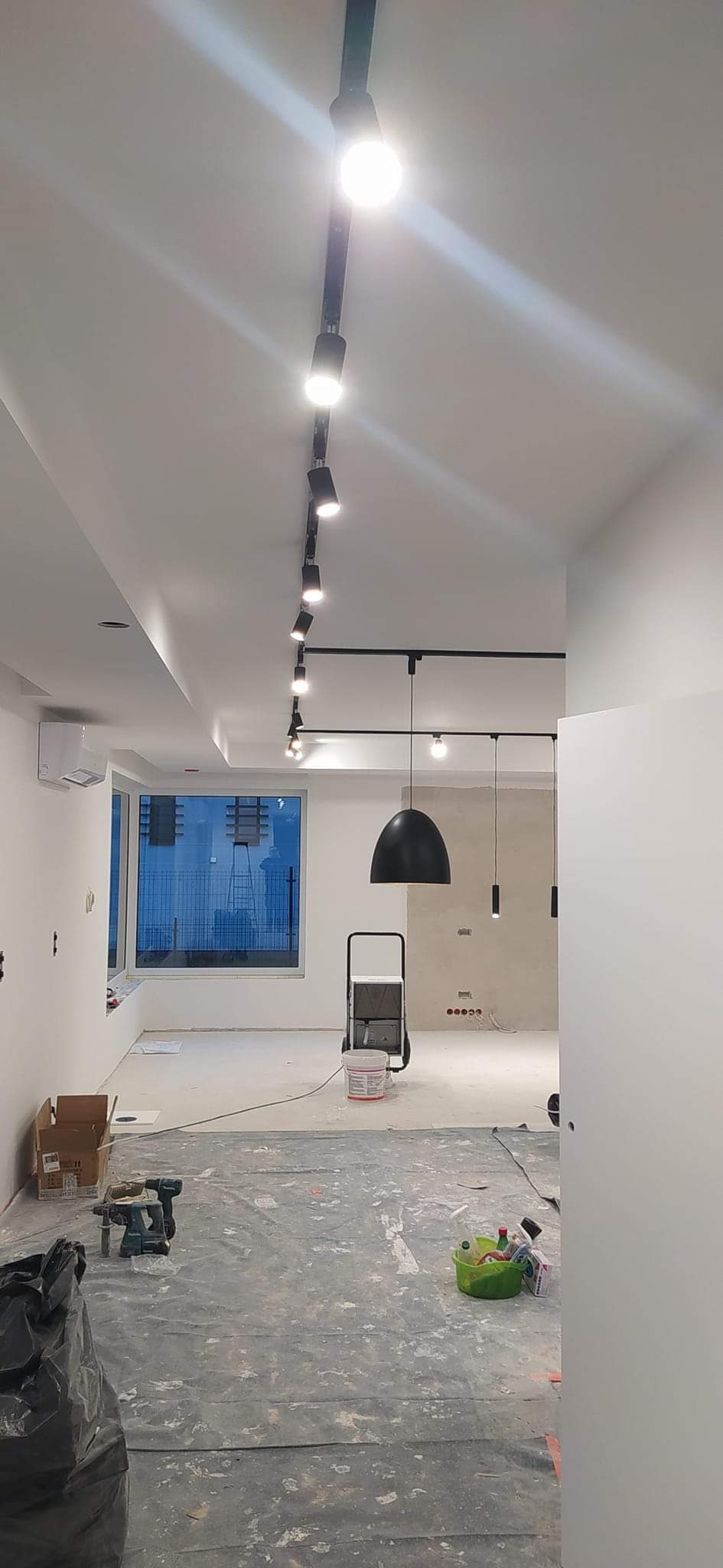 Wnętrze w trakcie remontu z zamontowanym systemem szynoprzewodów z reflektorami punktowymi i lampami wiszącymi, widoczny sprzęt budowlany i zabezpieczenia podłogi.