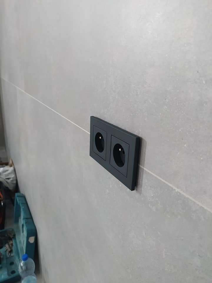Podwójne, czarne gniazdko elektryczne zamontowane na szarej, płytkowej ścianie z widoczną fugą i fragmentem skrzynki narzędziowej w tle.
