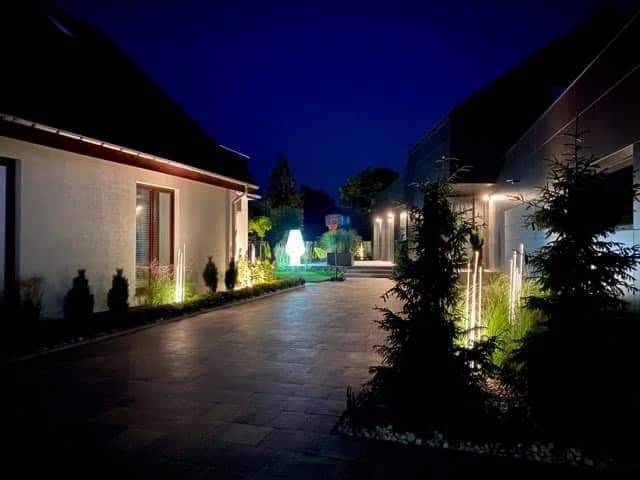 Oświetlona posesja nocą: nowoczesny dom z oświetleniem elewacyjnym i ogrodowym, widoczne lampy wzdłuż ścieżki i podświetlone krzewy.