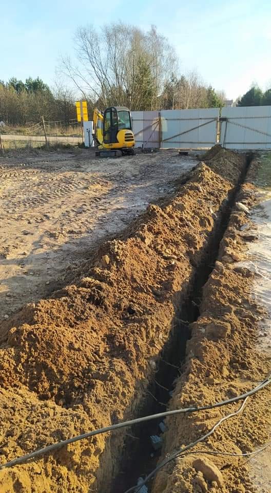 Wykopany rów w ziemi z widocznymi kablami wewnątrz, na drugim planie mała koparka i ogrodzenie.