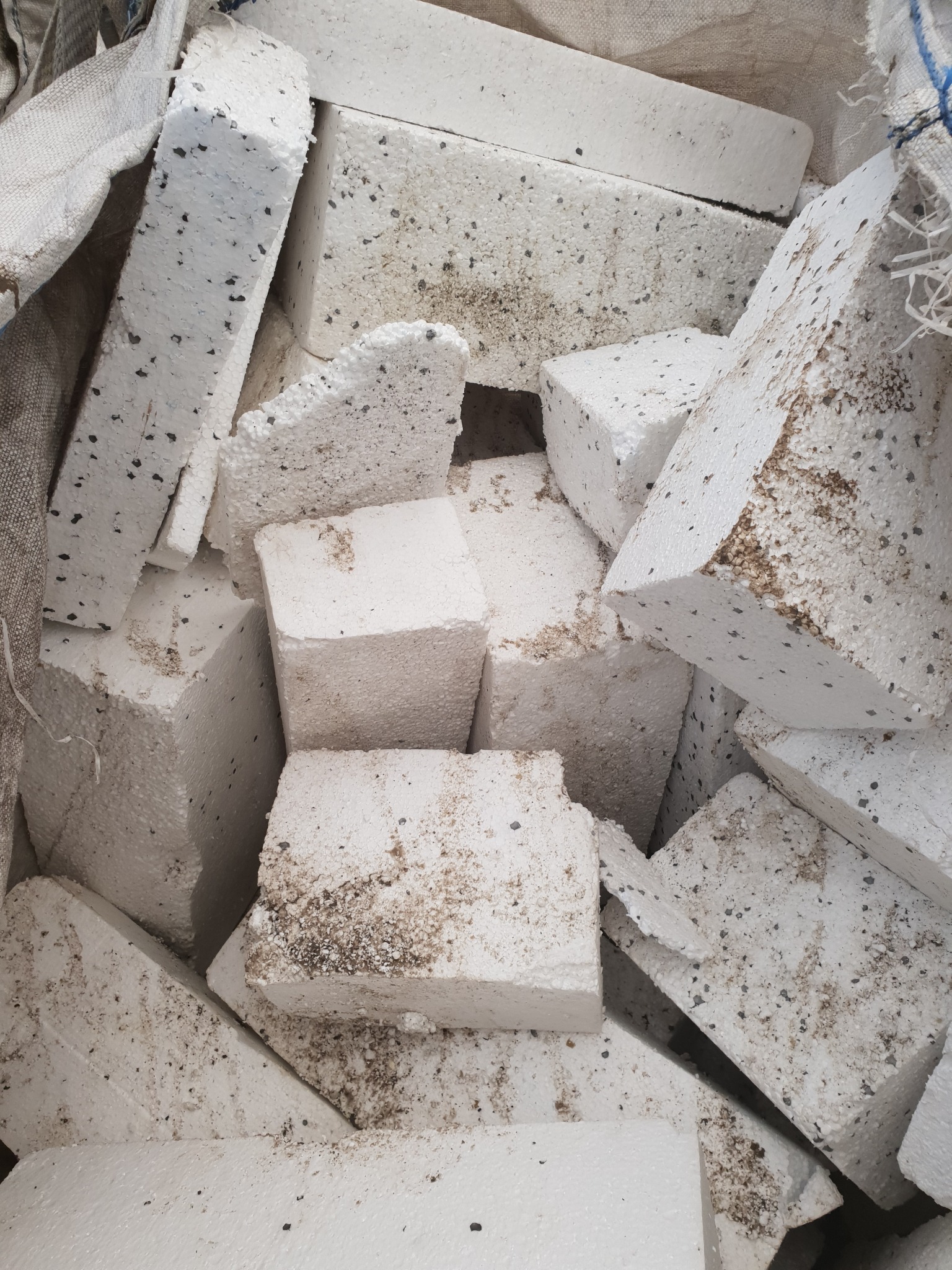 Kawałki brudnego styropianu w worku jutowym, widoczne ciemne plamy i ślady ziemi na powierzchni.