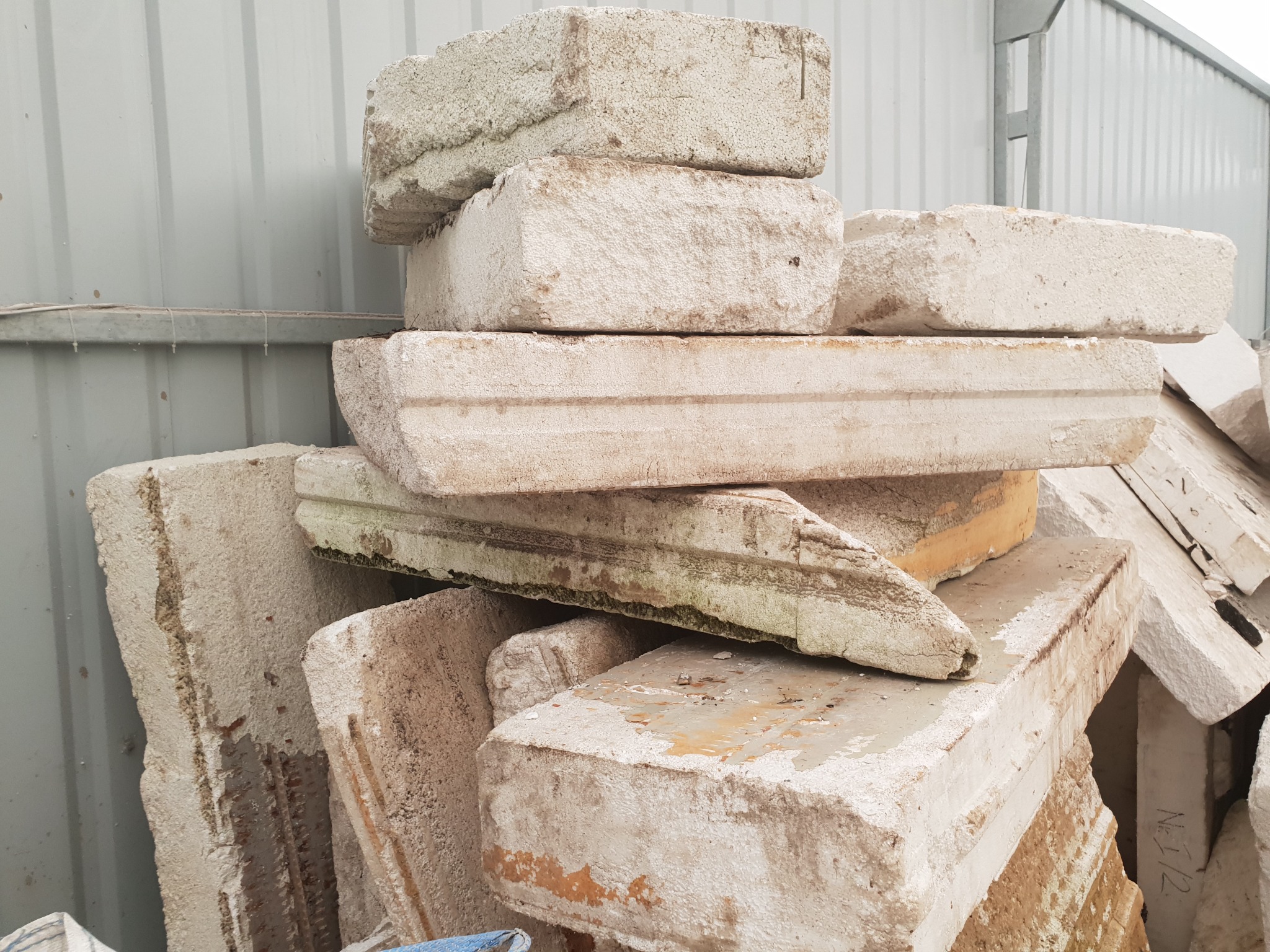 Stos białych, używanych płyt ociepleniowych z widocznymi śladami zabrudzeń i uszkodzeń, ułożonych na tle blaszanej ściany.