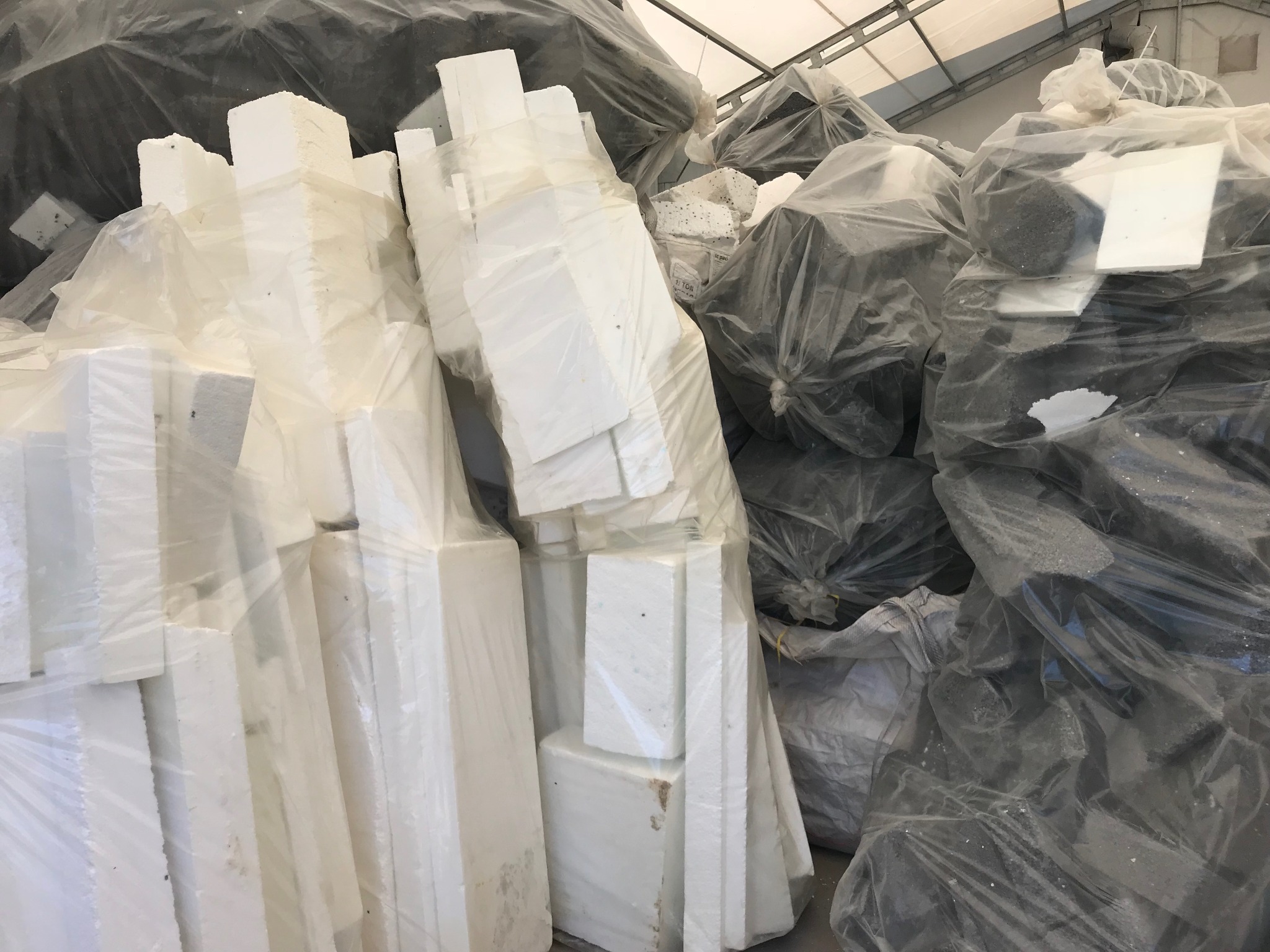 Skladowisko pakietów białych i szarych materiałów ociepleniowych w przezroczystych foliach, składowanych pod zadaszeniem.