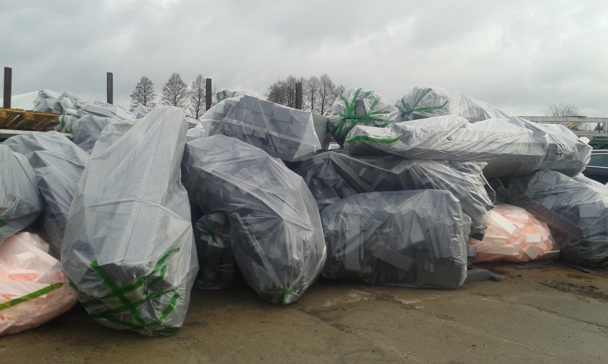 Stos pakietów materiałów izolacyjnych zabezpieczonych folią i zieloną taśmą na zewnątrz, widoczne elementy palet i drzewa w tle.