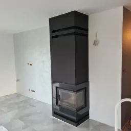 Nowoczesny, czarny kominek narożny z szybą, osadzony w wykończonym pomieszczeniu z jasnymi ścianami i błyszczącą, szarą podłogą z płytek.