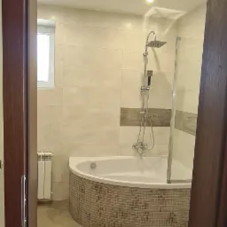 Wykończona łazienka z narożną wanną obudowaną mozaiką, beżowe płytki na ścianach i podłodze, widok zza otwartych, ciemnych drzwi.