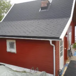 Czerwony budynek z czarnym dachem z gontów bitumicznych, okno zabezpieczone folią malarską, widoczny fragment rynny i komina.