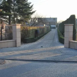 Wjazd do posesji z brukowaną drogą, ozdobnymi słupkami z piaskowca i zadbanym żywopłotem po bokach.