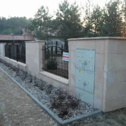Ogrodzenie z kamiennymi słupkami i metalowymi przęsłami, obok skrzynka elektryczna w kamiennej obudowie, widoczne krzewy i kamyczki dekoracyjne.