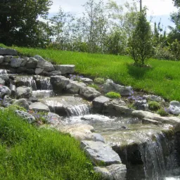 Kaskada wodna w ogrodzie, zbudowana z szarych kamieni, z roślinami i kwiatami pomiędzy nimi, na tle zielonego trawnika i drzew.