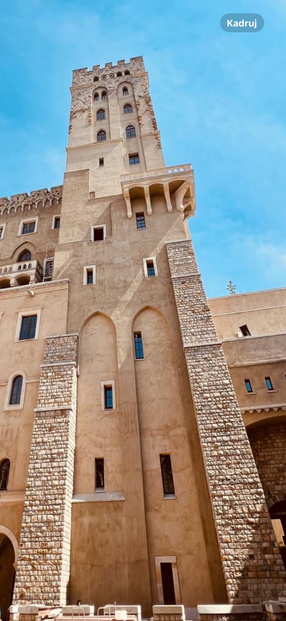 Fasada budynku z wysoką wieżą, z elementami dekoracyjnymi z kamienia, uchwycona pod kątem z dołu, ukazująca architekturę i detale wykończenia.