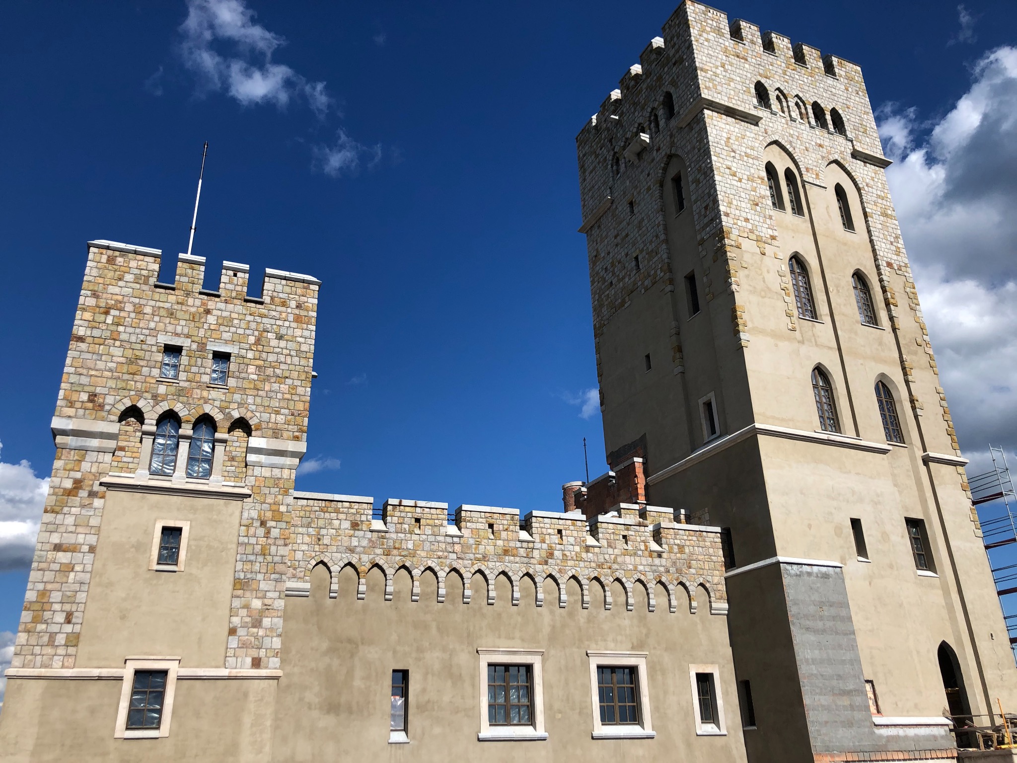 Fasada zamku z kamiennymi elementami i wieżą na tle błękitnego nieba z chmurami, widoczne rusztowania.