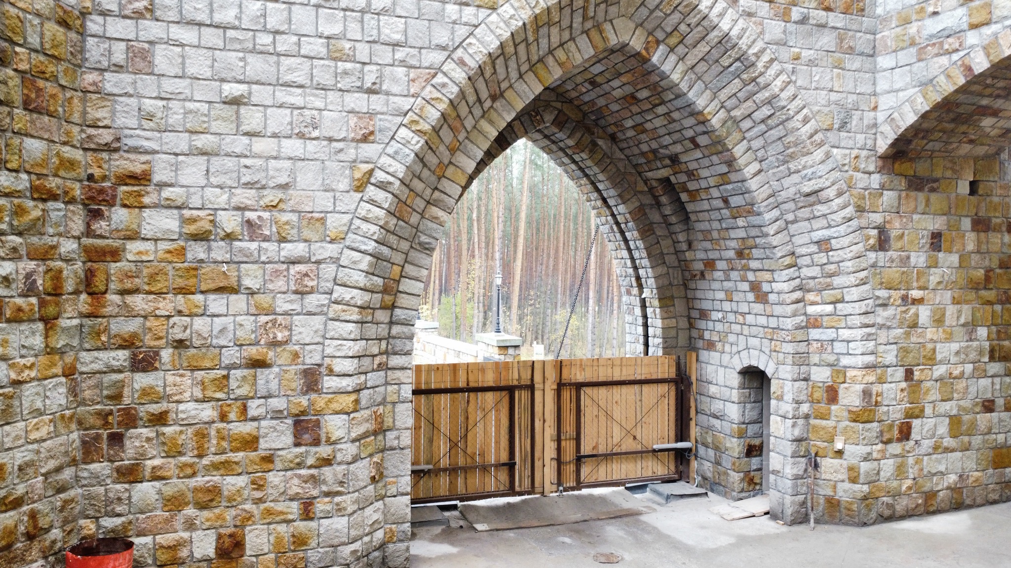 Kamienny łuk z drewnianą bramą w tle lasu, mur z nieregularnych kamieni w odcieniach szarości i beżu, widoczny fragment drugiego łuku i małe okienko w murze.