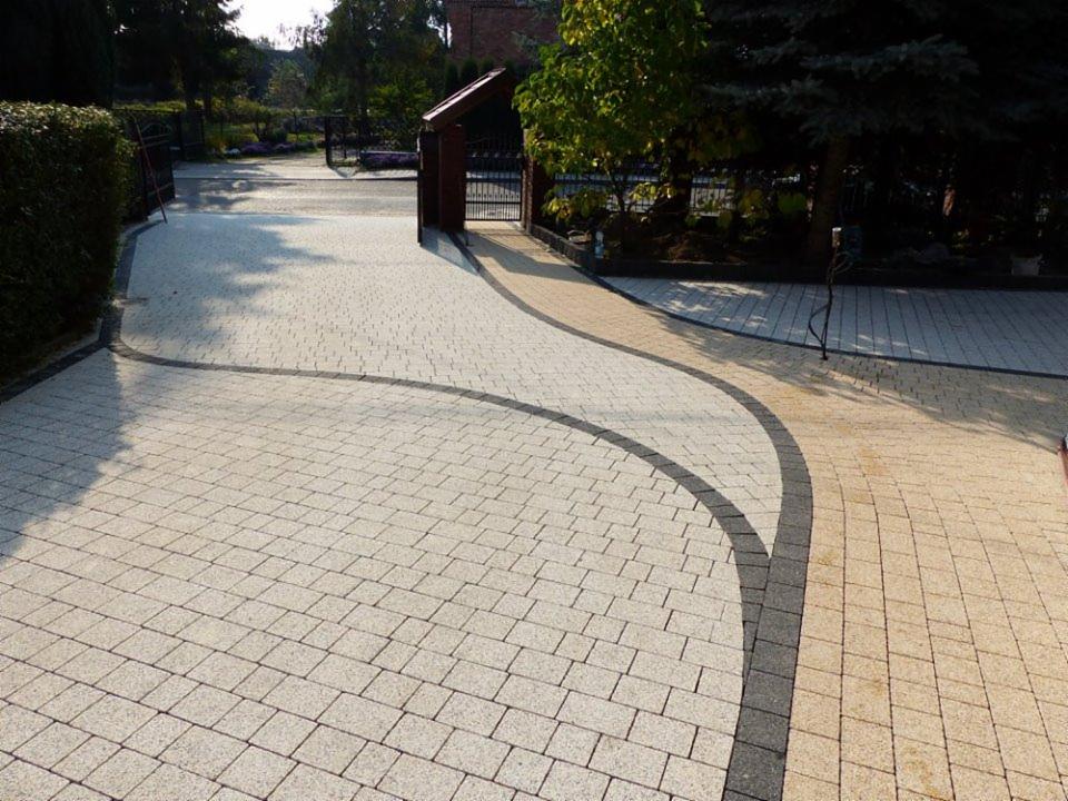 Ułożona kostka brukowa w odcieniach szarości i beżu tworząca ścieżkę w ogrodzie, z ciemnoszarą obwódką i bramą w tle.