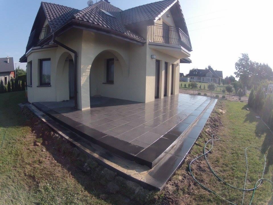 Nowoczesny taras z ciemnej kostki na dwóch poziomach przy domu z balkonem, widok z perspektywy żabiej z lekkim odblaskiem na powierzchni kostki.