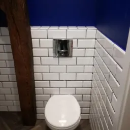 Nowoczesna toaleta z białymi, prostokątnymi płytkami ułożonymi w cegiełkę, kontrastującymi z ciemnoniebieską ścianą i rustykalną, drewnianą belką. Na podłodze parkiet ułożony w jodełkę.