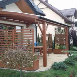 Zadaszony taras z drewnianą konstrukcją, ażurowymi panelami i donicami, wyłożony jasną kostką brukową, otoczony zielenią ogrodową.