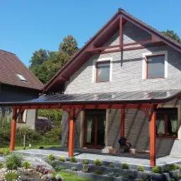 Nowoczesna pergola z drewnianą konstrukcją i przezroczystym zadaszeniem, przylegająca do domu z szarą elewacją, widok na zadbany ogród z kamiennymi rabatami i pracownika podczas montażu.