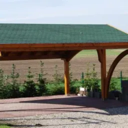 Drewniana konstrukcja z zielonym dachem, prawdopodobnie wiata garażowa, z łukowym wsparciem, przed nią drewniana drabinka ogrodowa, w tle pole i trawa.