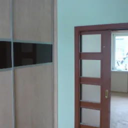 Beżowa szafa przesuwna z ciemnymi pasami i brązowe drzwi z szybkami w odnowionym pokoju z grzejnikiem pod oknem.