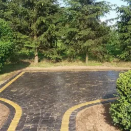 Czarna kostka brukowa ułożona w łukowy wzór z żółtymi obrzeżami, otoczona zielenią drzew i krzewów, tworząca parking lub plac.