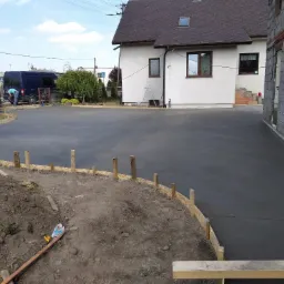 Świeżo wylany, gładki jastrych na podjeździe przed domem jednorodzinnym, z widocznym szalunkiem z drewnianych desek i pomocnikiem przy samochodzie w tle.