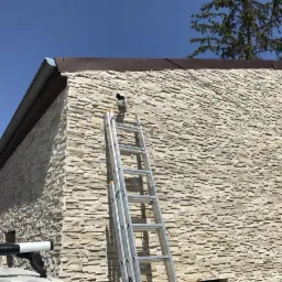 Kamera monitoringu zamontowana na elewacji budynku wykończonej kamieniem dekoracyjnym, widoczna aluminiowa drabina oparta o ścianę.