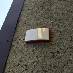 Zewnętrzna sygnalizacja alarmowa, biało-czerwona, zamontowana na szorstkiej, betonowej ścianie budynku, obok metalowej rynny.