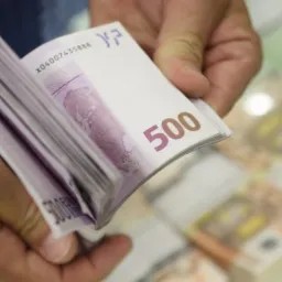 Stos 500-euro banknotów trzymanych w dłoniach, z widocznym numerem seryjnym na jednym z banknotów, w tle rozmyte stosy banknotów.
