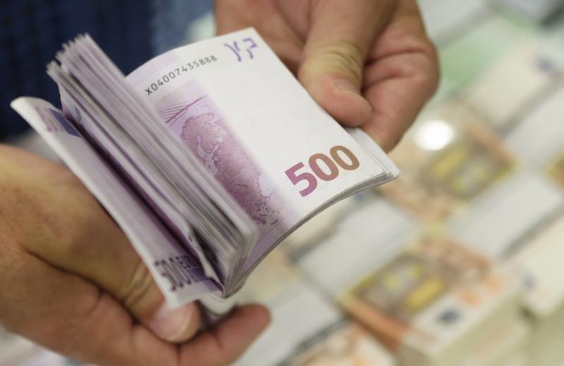 Stos 500-euro banknotów trzymanych w dłoniach, z widocznym numerem seryjnym na jednym z banknotów, w tle rozmyte stosy banknotów.