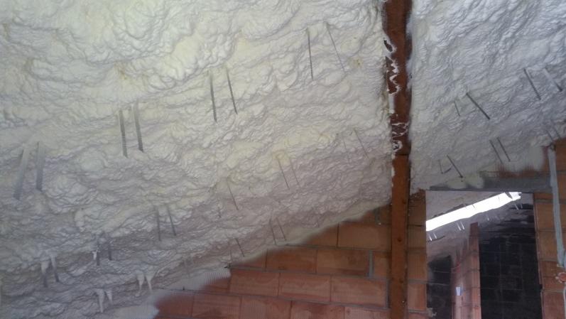 Poddasze w trakcie izolacji, pokryte białą pianą z widocznymi metalowymi szpilkami mocującymi, ceglana ściana w dolnej części kadru.