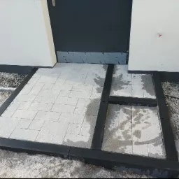 Nowo ułożony, jasnoszary chodnik z kostki brukowej obramowany czarnym, błyszczącym krawężnikiem, prowadzący do drzwi wejściowych budynku. Część kostki jest jeszcze mokra, a na obrzeżach widać gruz...