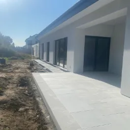 Jasnoszary taras z płyt betonowych przylegający do nowoczesnego domu z ciemnymi oknami, widoczny fragment ogrodu z przygotowanym podłożem.