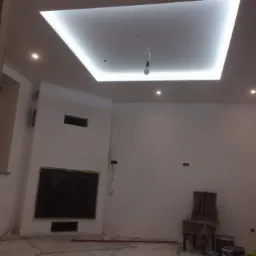 Nowoczesny sufit podwieszany z oświetleniem LED w kształcie kwadratu, widok na pokój w trakcie remontu, z zabezpieczonym miejscem na telewizor, krzesłem pod folią i drabiną opartą o ścianę.