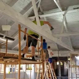 Trzech mężczyzn na rusztowaniach i drabinie maluje na biało drewniany sufit z widoczną konstrukcją nośną w jasnym pomieszczeniu z oknami, podłoga zabezpieczona folią malarską.