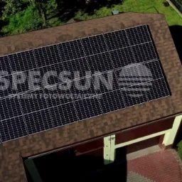 Widok z góry na panele słoneczne z logo firmy na dachu domu z brązową dachówką bitumiczną, obok garaż i trawnik.