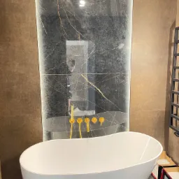 Nowoczesna łazienka z wolnostojącą białą wanną, złotą armaturą i ścianą wyłożoną płytkami imitującymi czarny marmur z złotymi żyłami, odbicie fotografa w tafli płytek.