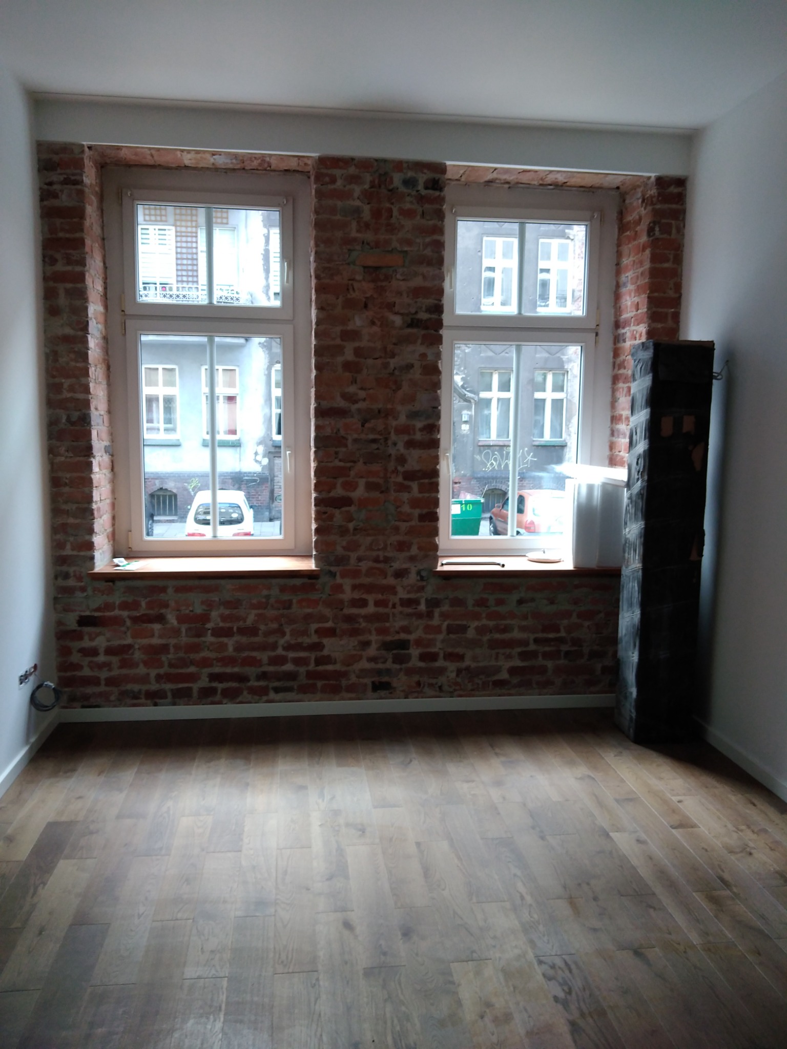 Wnętrze pokoju z dwiema oknami z ceglanym obramowaniem, nowa drewniana podłoga ułożona w jodełkę, widoczny fragment grzejnika obok okna, ściany białe.