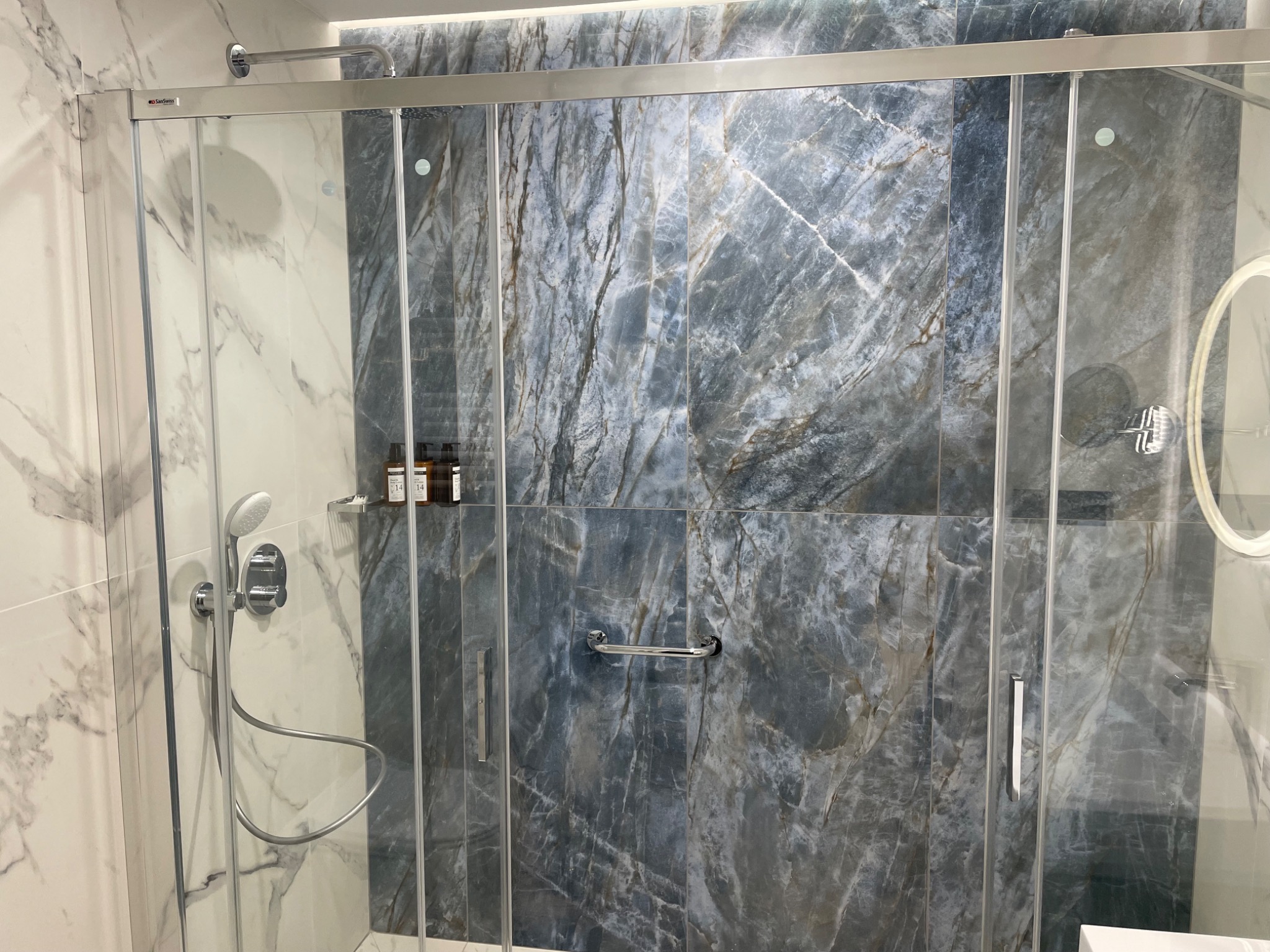 Szklana kabina prysznicowa z przesuwnymi drzwiami, wykończona płytkami imitującymi marmur w odcieniach szarości i bieli, z chromowaną armaturą i półką na kosmetyki.