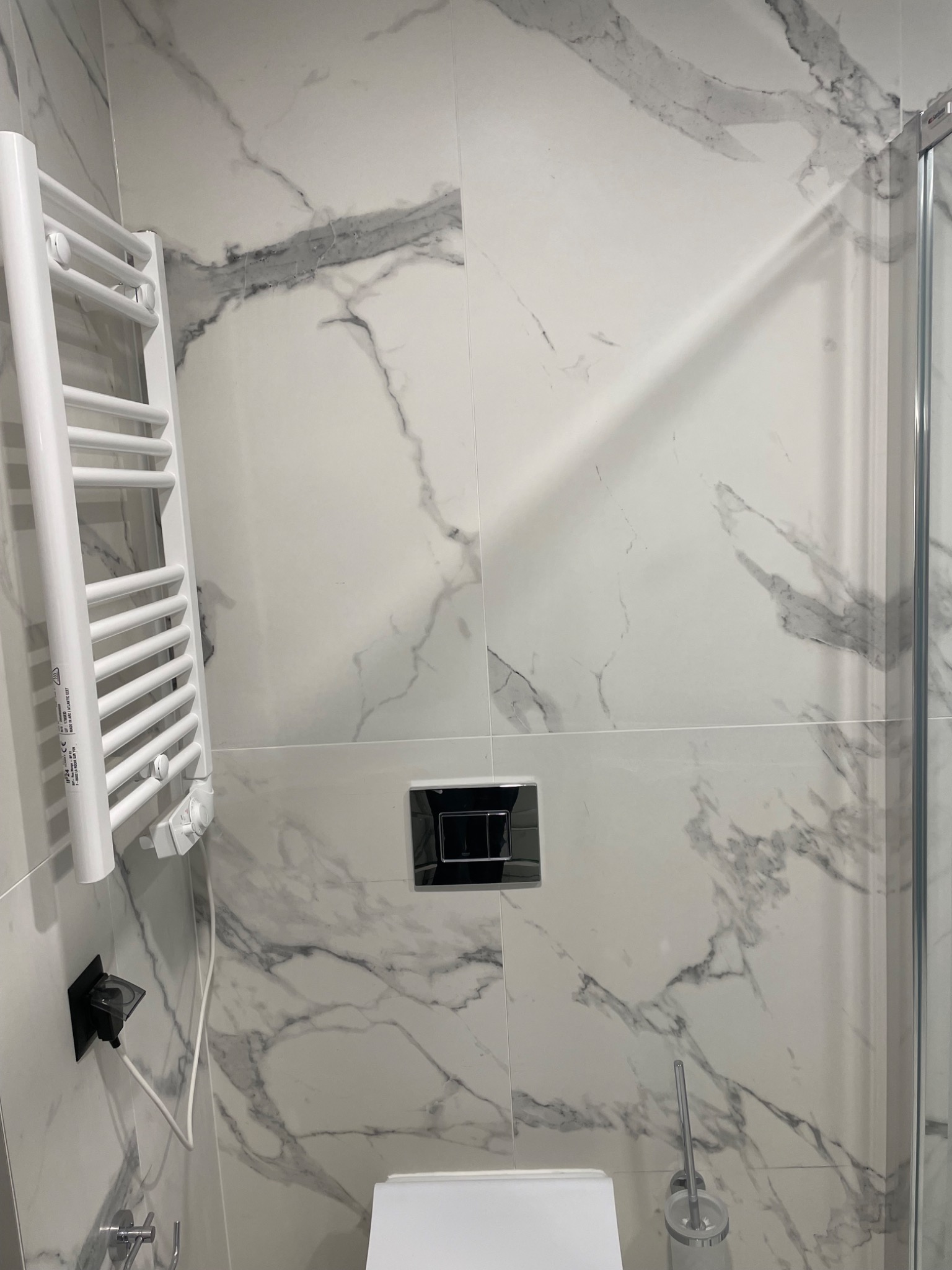 Wykończona łazienka z białymi płytkami imitującymi marmur, widoczny grzejnik drabinkowy, toaleta i fragment kabiny prysznicowej.