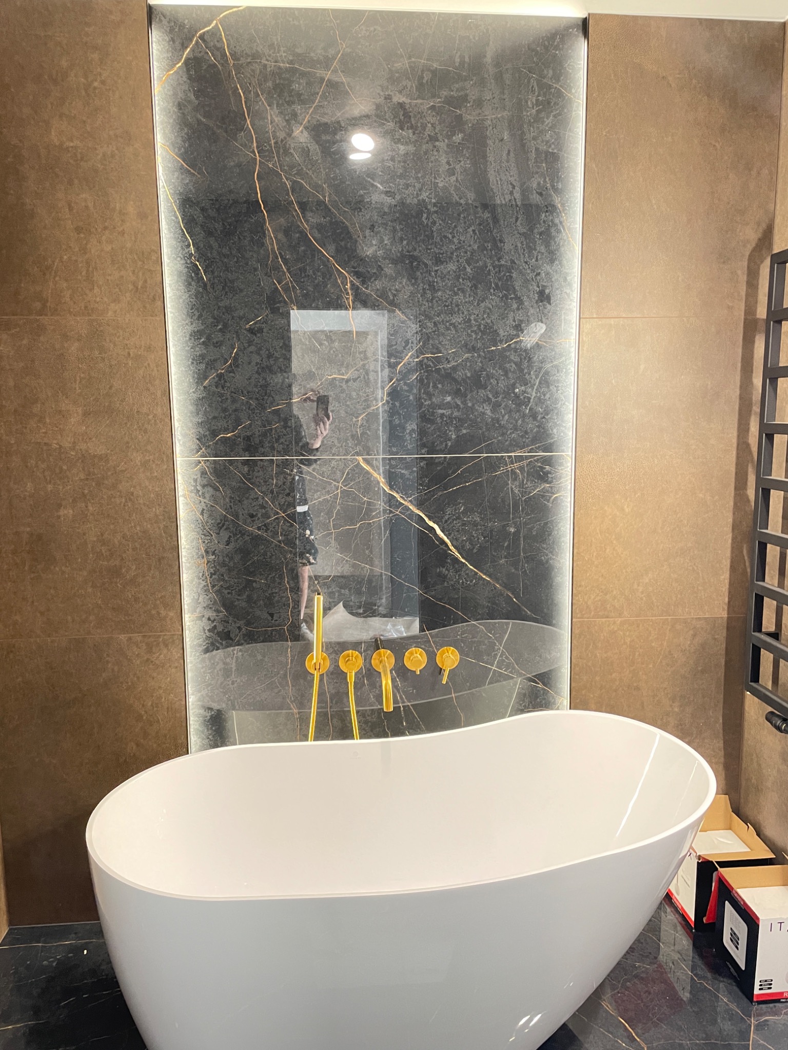 Nowoczesna łazienka z wolnostojącą białą wanną, złotą armaturą i ścianą wyłożoną płytkami imitującymi czarny marmur z złotymi żyłami, odbicie fotografa w tafli płytek.