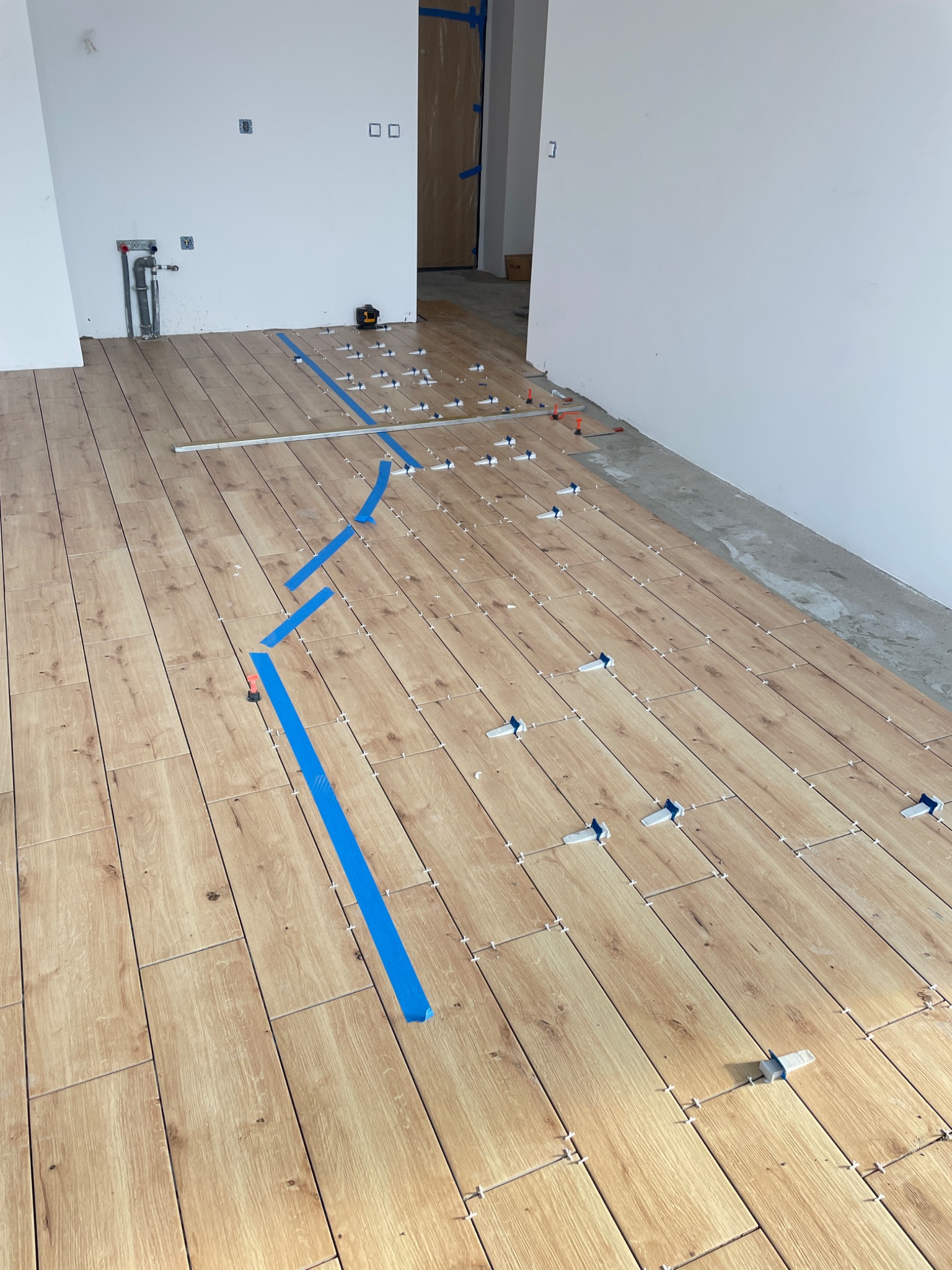 Układanie podłogi z płytek imitujących drewno, z użyciem poziomicy laserowej, niebieskiej taśmy maskującej i systemu poziomowania płytek, widoczne białe ściany i instalacja wodna.