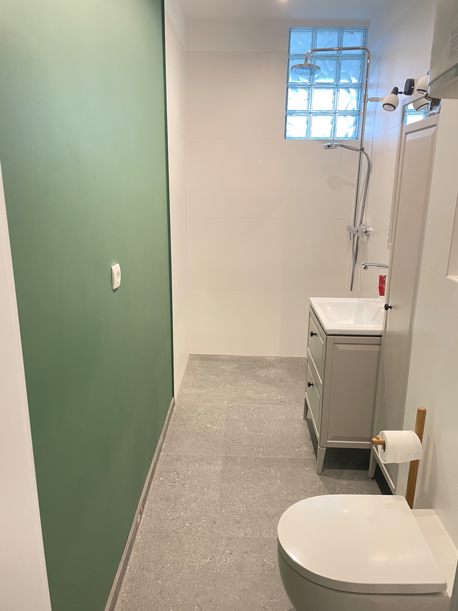 Nowoczesna łazienka z zieloną ścianą, szarą podłogą, umywalką z szafką i toaletą, oświetlona przez luksfery w strefie prysznica.