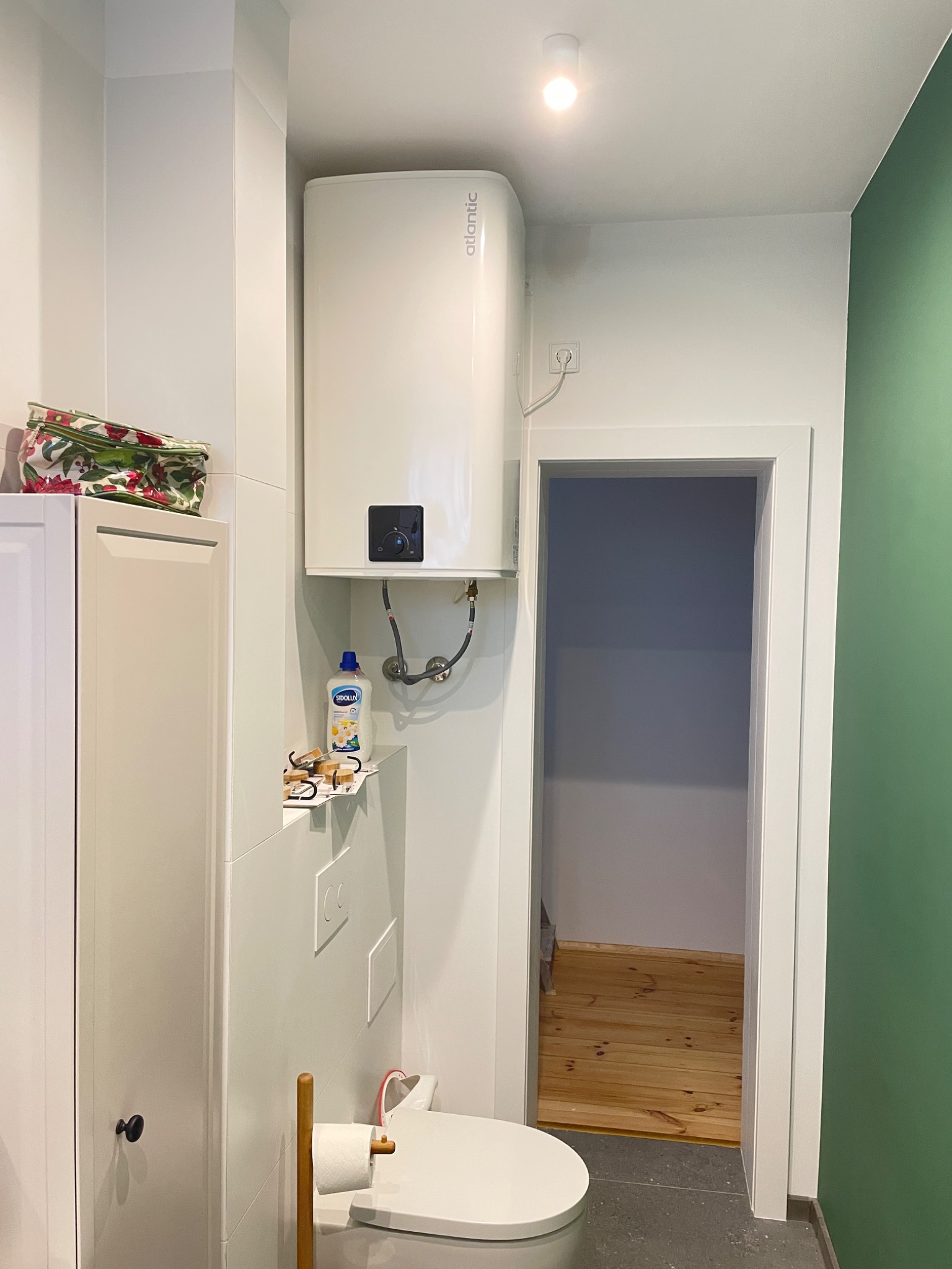 Biała łazienka z zamontowanym bojlerem Atlantic, toaletą, szafką, półką z płynem do czyszczenia i akcesoriami, oraz widokiem na drewnianą podłogę w sąsiednim pomieszczeniu przez otwarte drzwi.
