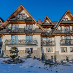 Elewacja pensjonatu w stylu g&oacute;ralskim z balkonami z kutymi balustradami, wykończona kamieniem, z widocznymi śladami śniegu.