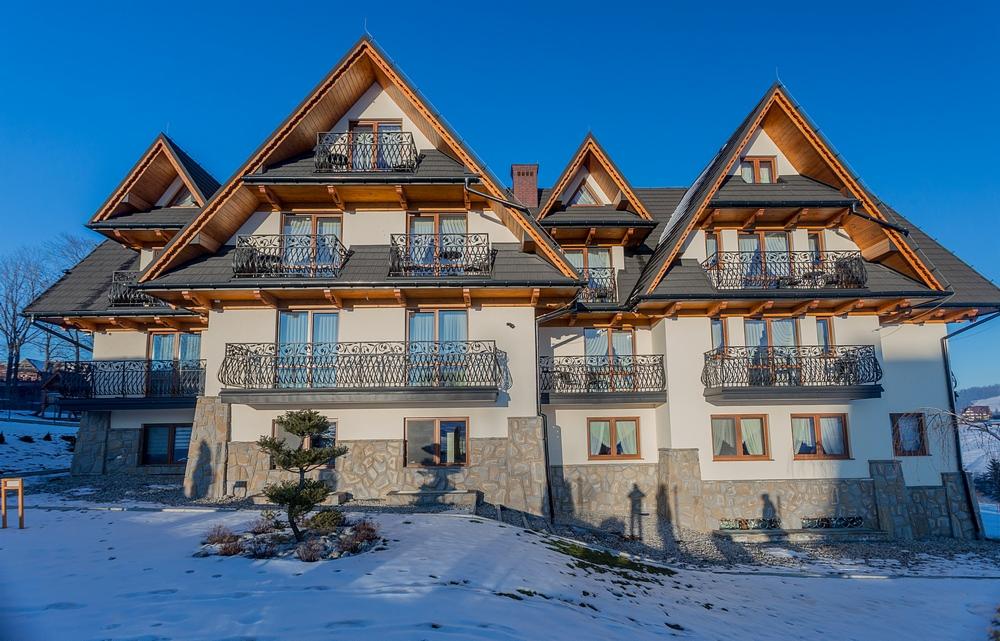Elewacja pensjonatu w stylu góralskim z balkonami z kutymi balustradami, wykończona kamieniem, z widocznymi śladami śniegu.