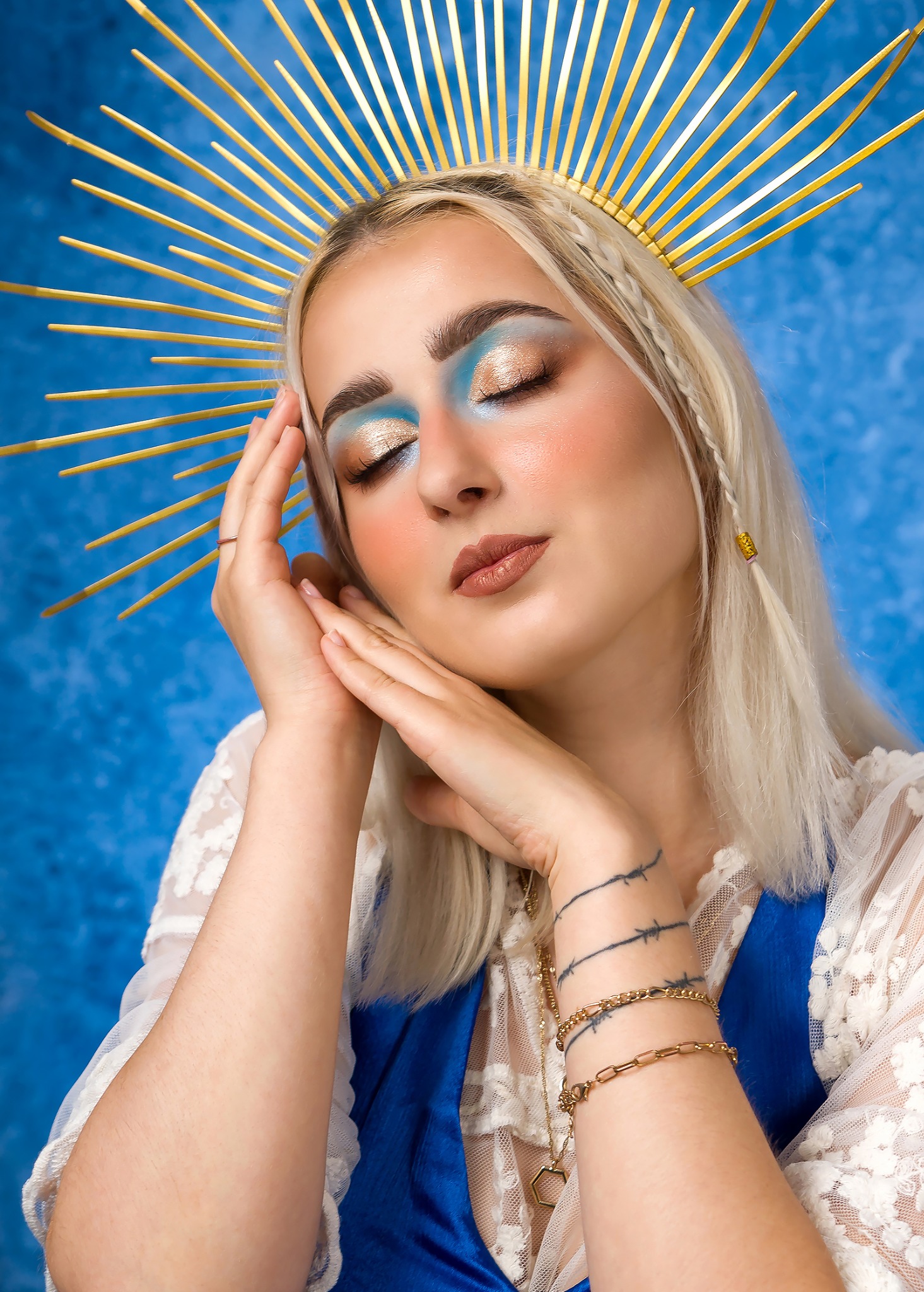 Portret kobiety z zamkniętymi oczami, w stylizowanej koronie z promieniami, z niebieskim cieniem na powiekach i delikatnym makijażem, na tle błękitnej, teksturowanej ściany.